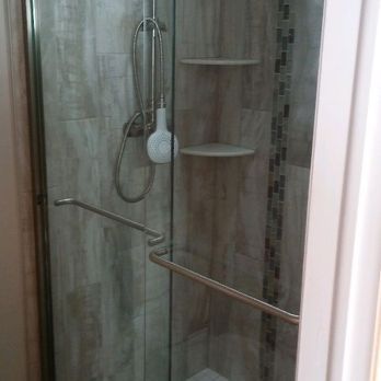 Small bathroom shower with glass door, handheld showerhead, corner shelf, and grab bars.