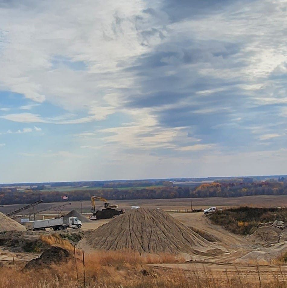 Wide rural construction site with excavator, dump truck, and dirt roads under a cloudy sky