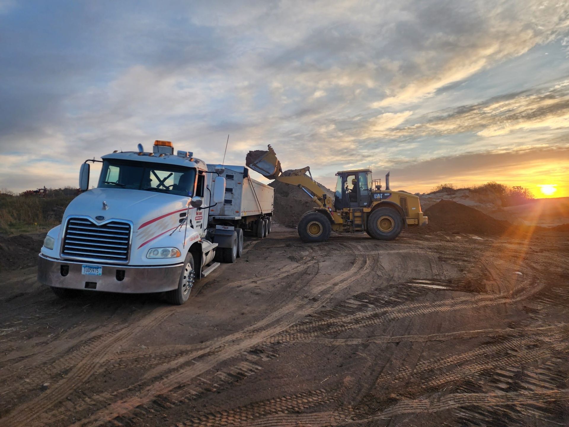 Construction trucks and loader on a dirt road at sunset, with a dump truck in the foreground.