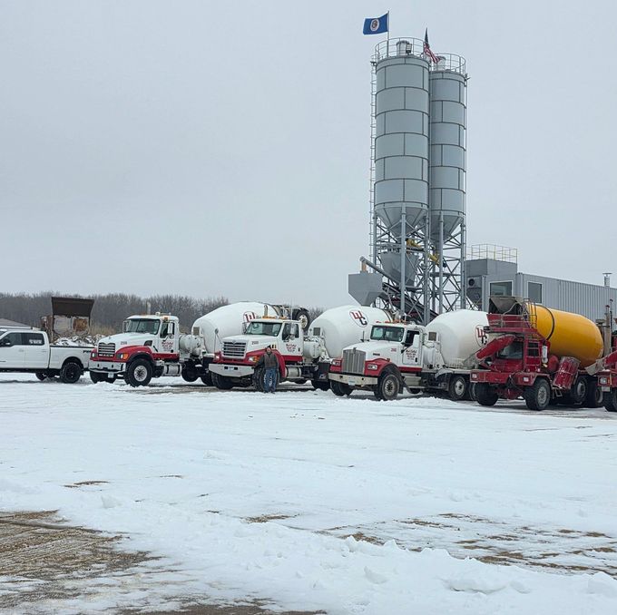 Cement plant with tall silos and several concrete mixer trucks parked on a snowy lot.