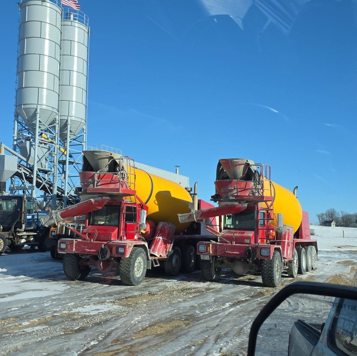 Two red cement mixers in a snowy industrial yard beside tall silos under a clear blue sky