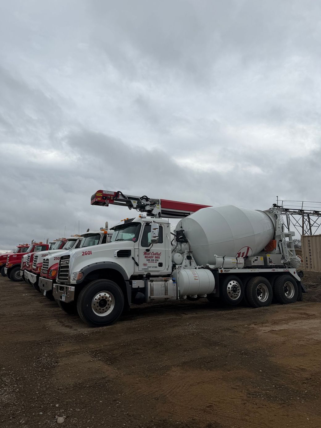 White concrete mixer truck with red crane parked on a muddy lot under cloudy skies