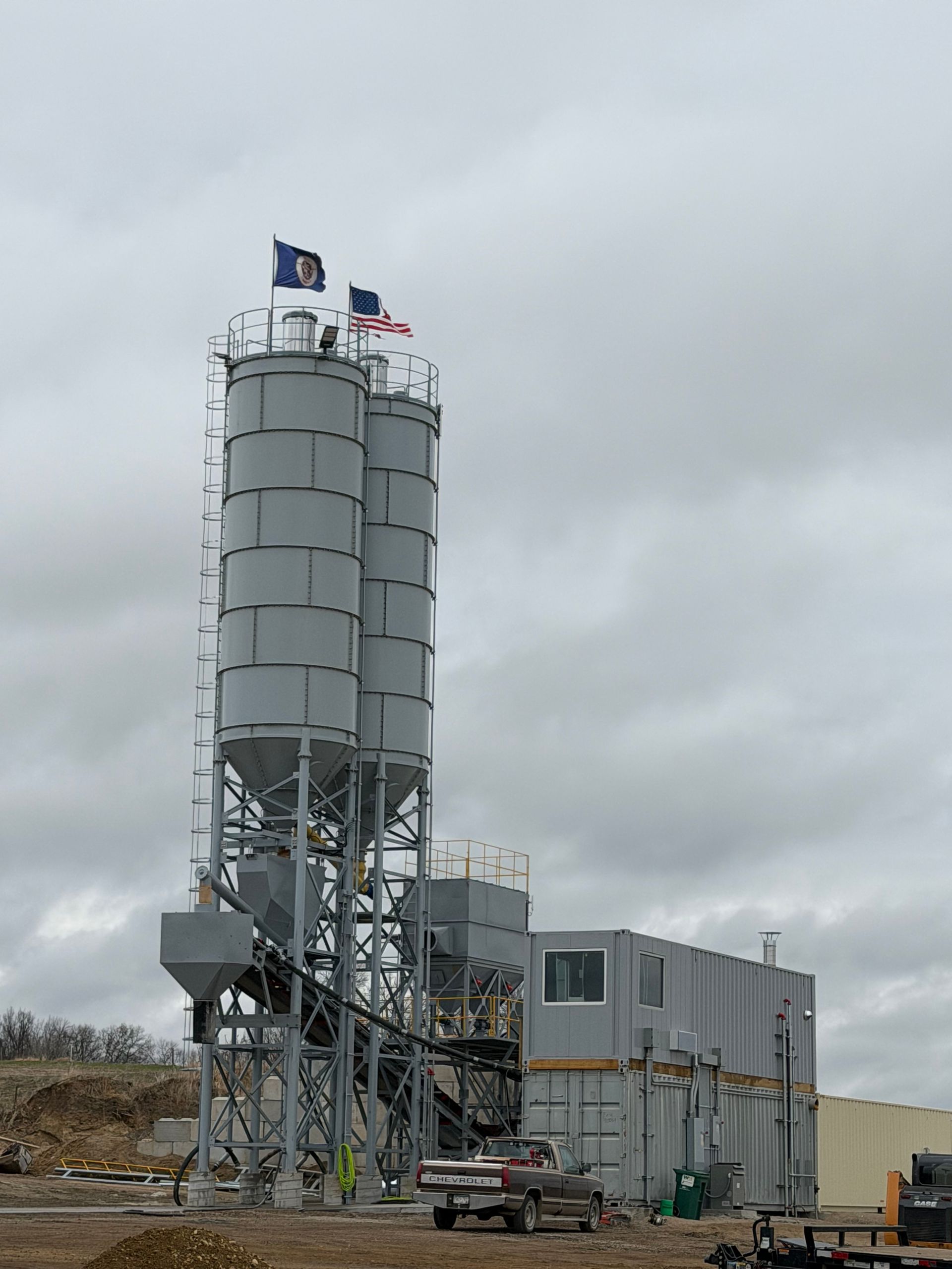 Industrial silo and processing building under a cloudy sky, with flags on top.