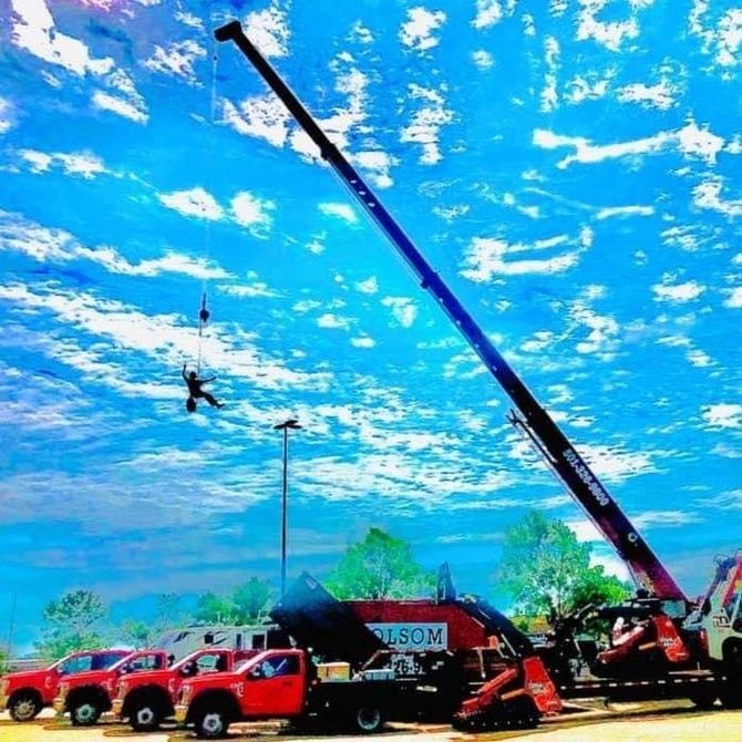 Crane lifting a hanging person above red trucks at an outdoor event under a bright blue sky