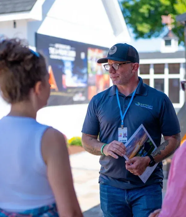Man in blue polo and cap speaking to attendees outdoors, holding brochures.