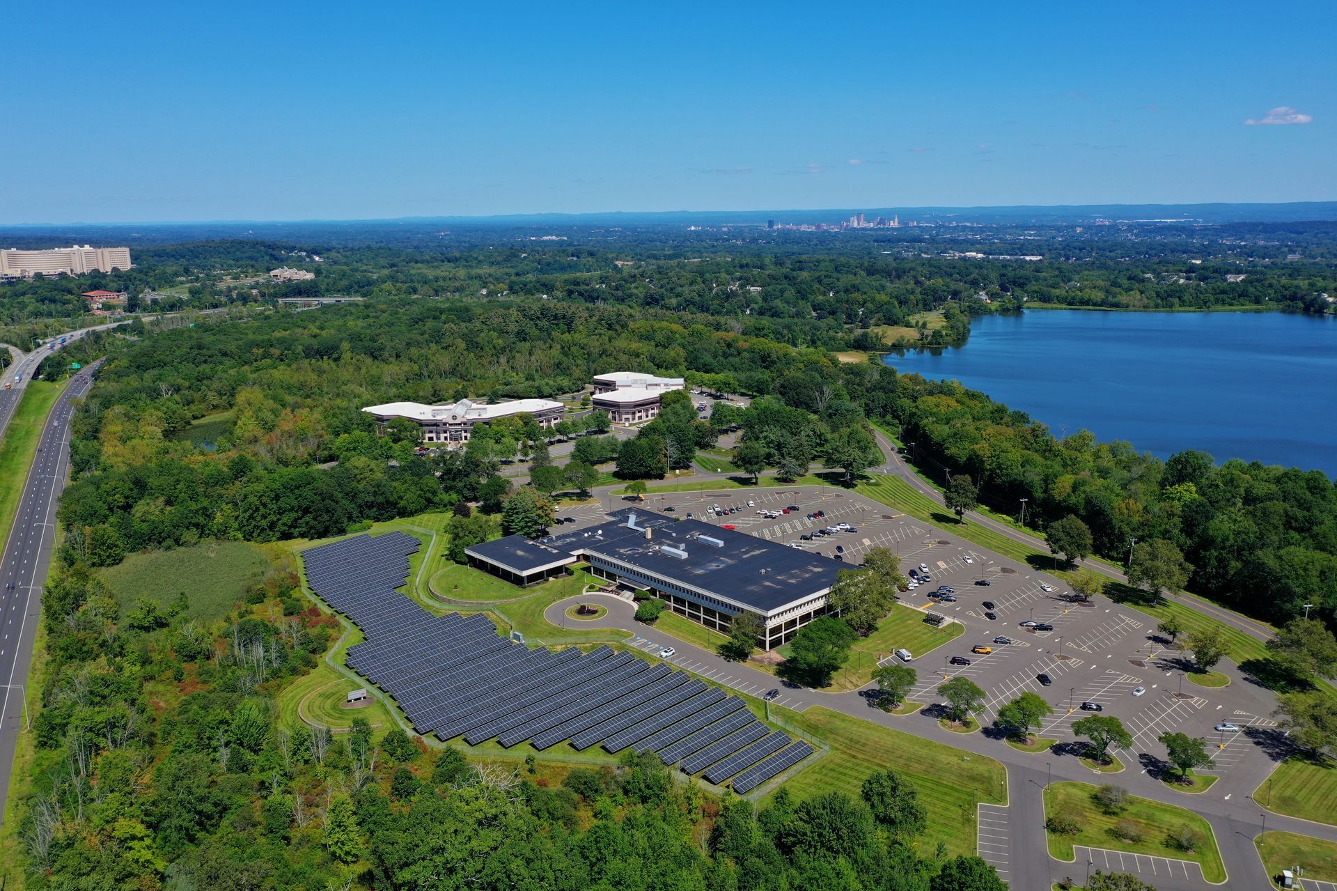 Aerial view of a solar panel farm beside a building, parking lot, and lake surrounded by green trees