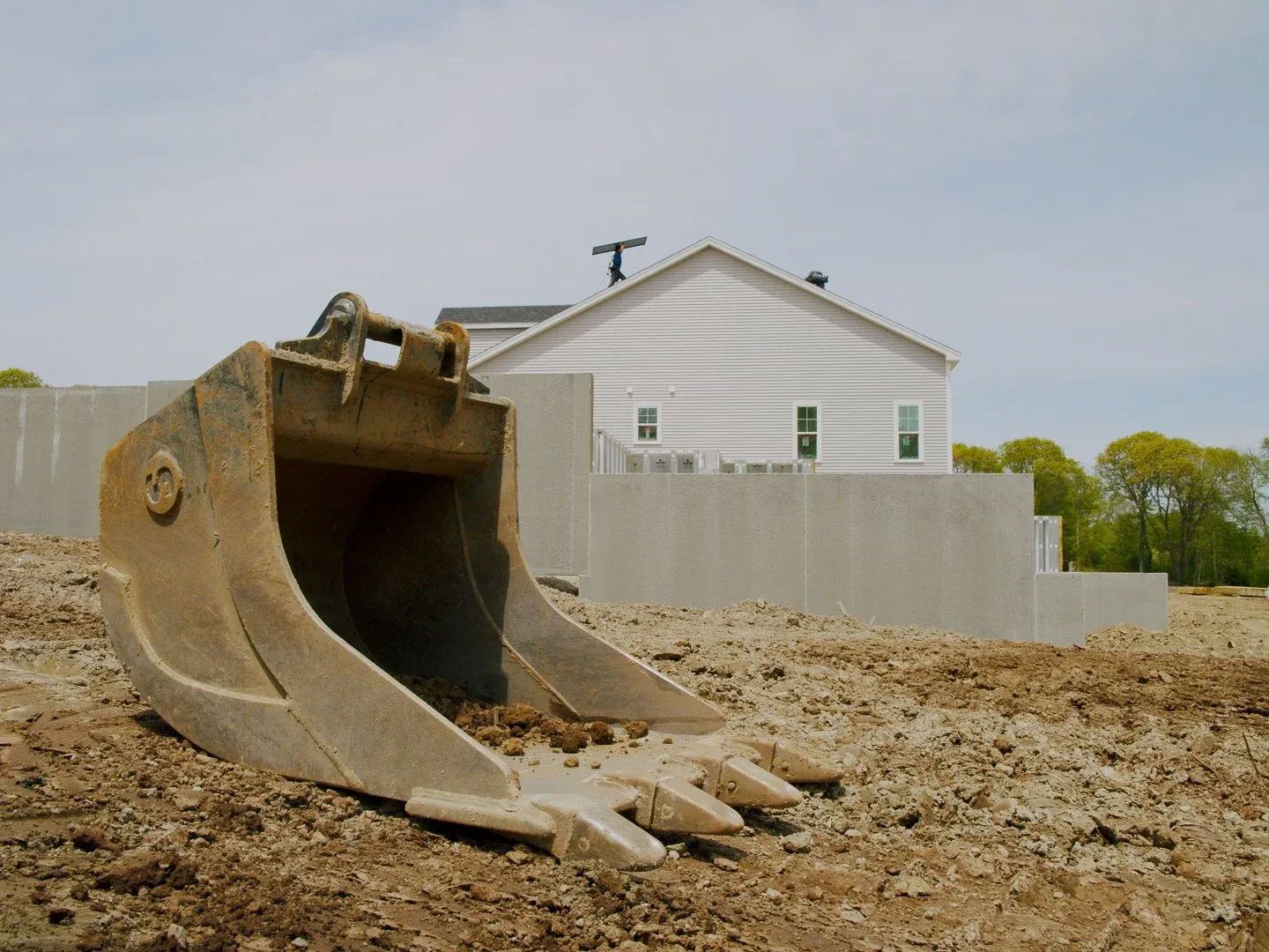 Excavator bucket in a dirt lot before a white house and gray fence