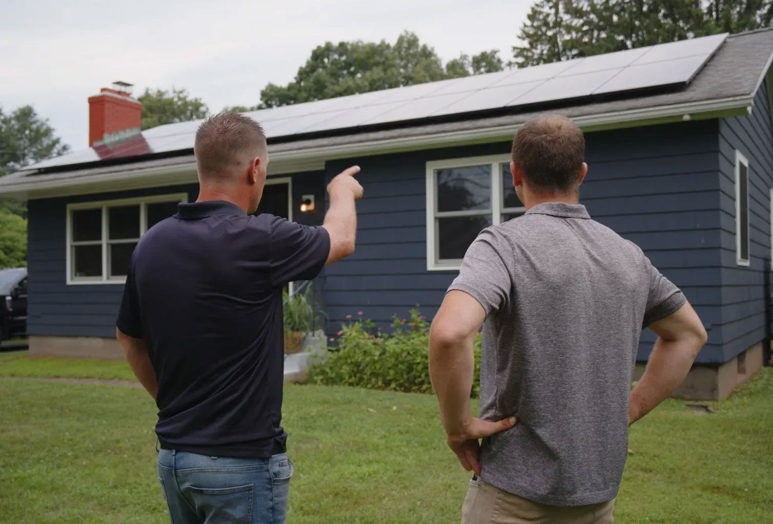Two men inspect a blue house with a roof solar panel, one pointing at the roof.
