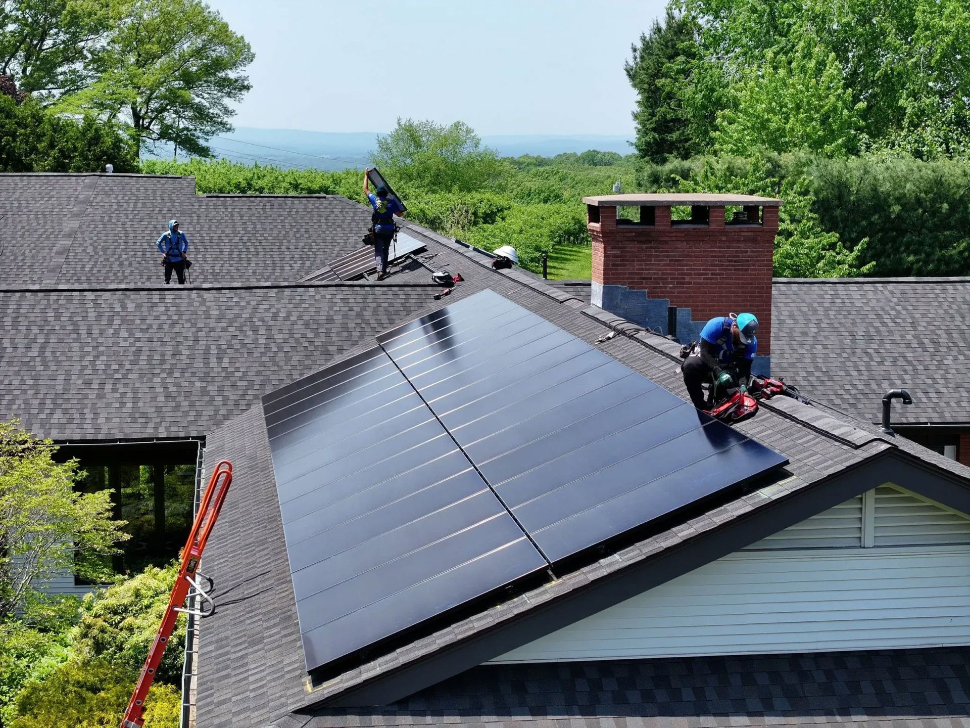 Workers installing solar panels on a house roof, with a wooded landscape in the background.