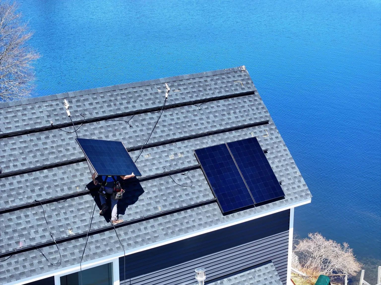 Worker on a gray shingled roof installing a dark blue solar panel in bright sunlight