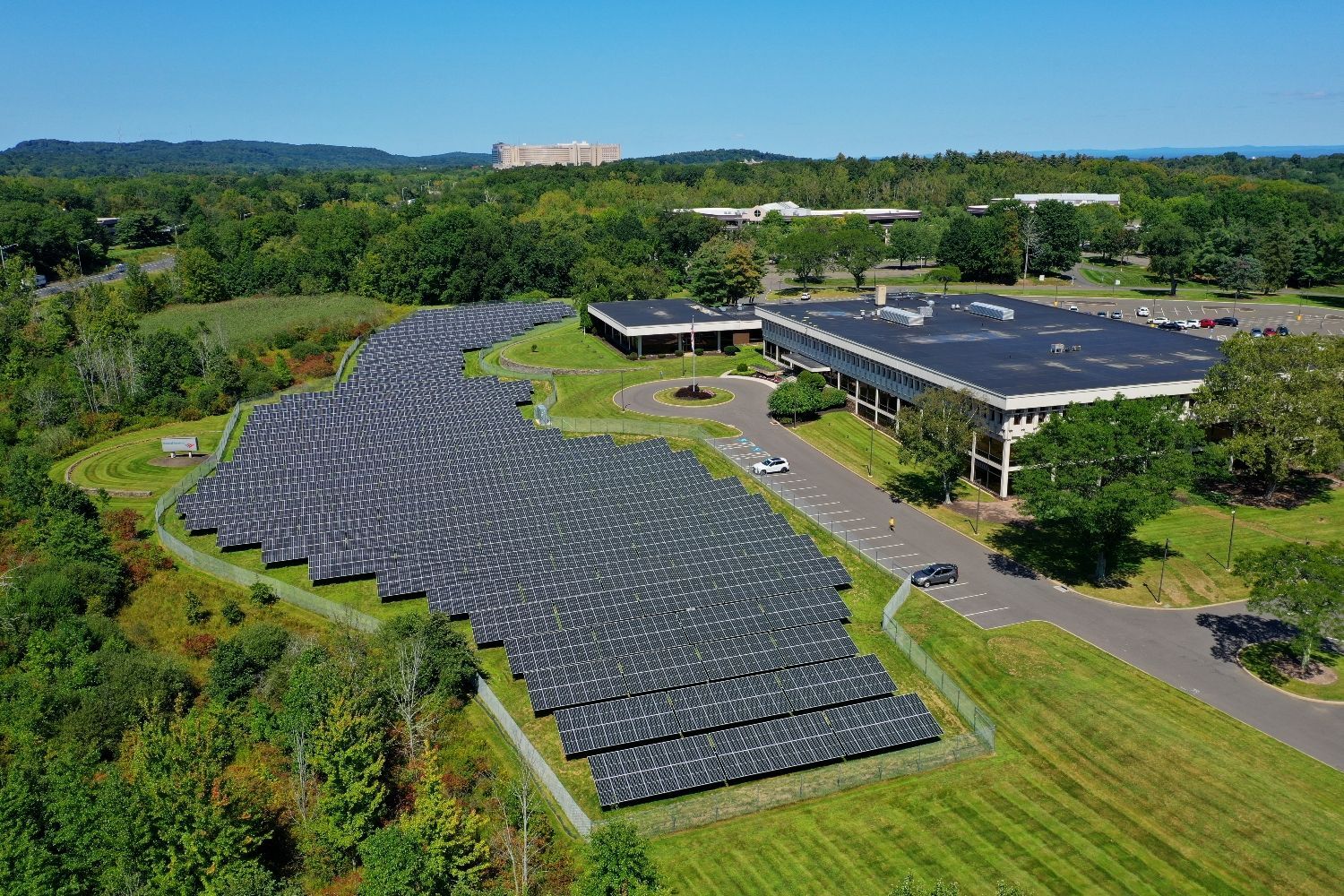 Aerial view of a large solar panel array beside industrial buildings and trees on a sunny day