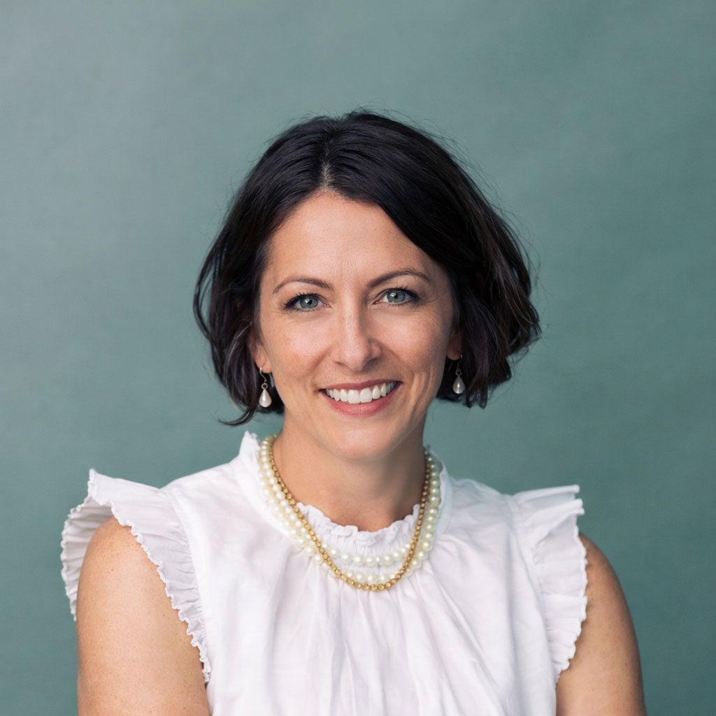 Smiling woman with short dark hair wearing a white ruffled blouse and pearl necklace against a gray background