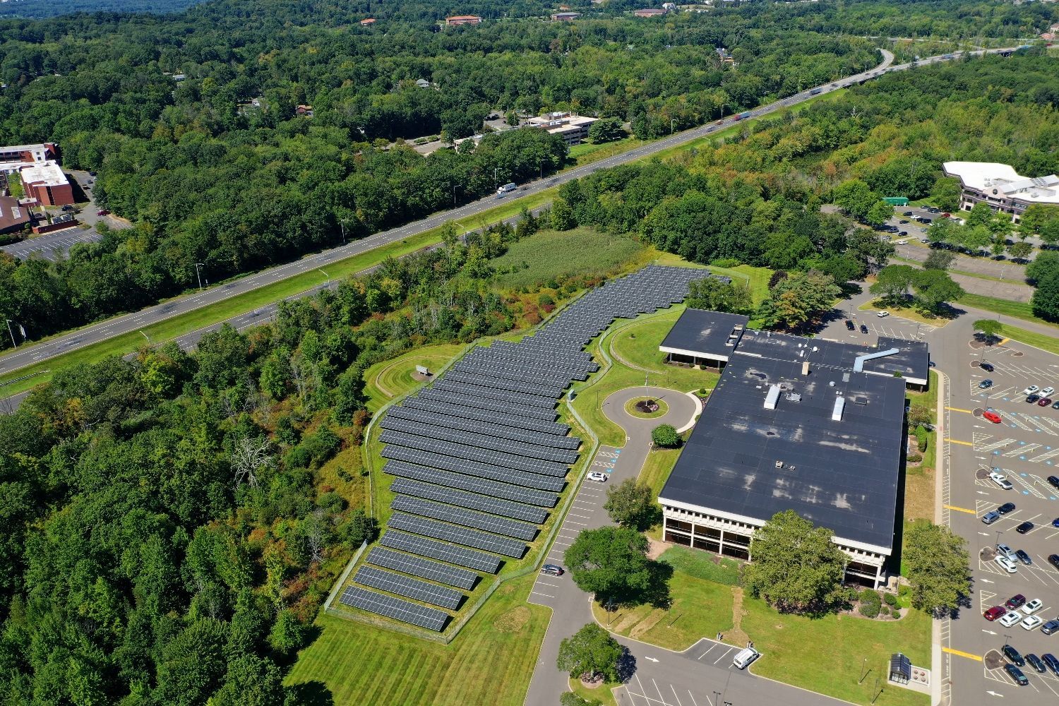Aerial view of a building with solar panels beside a wooded area and parking lot.