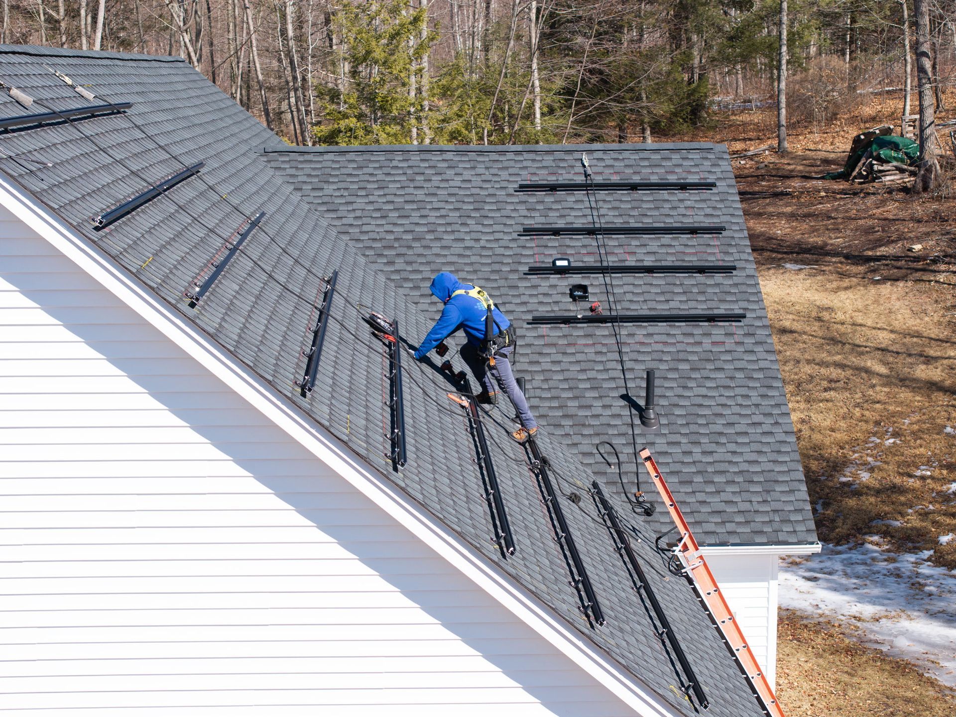 Worker in blue jacket installing black roofing panels on a steep gray roof with safety harness.