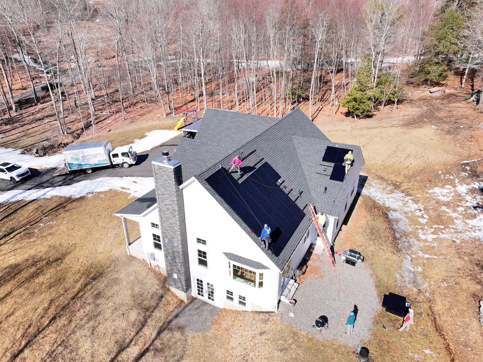 Aerial view of a white house with a dark roof, parked vehicles, and people in a wooded yard with patchy snow.