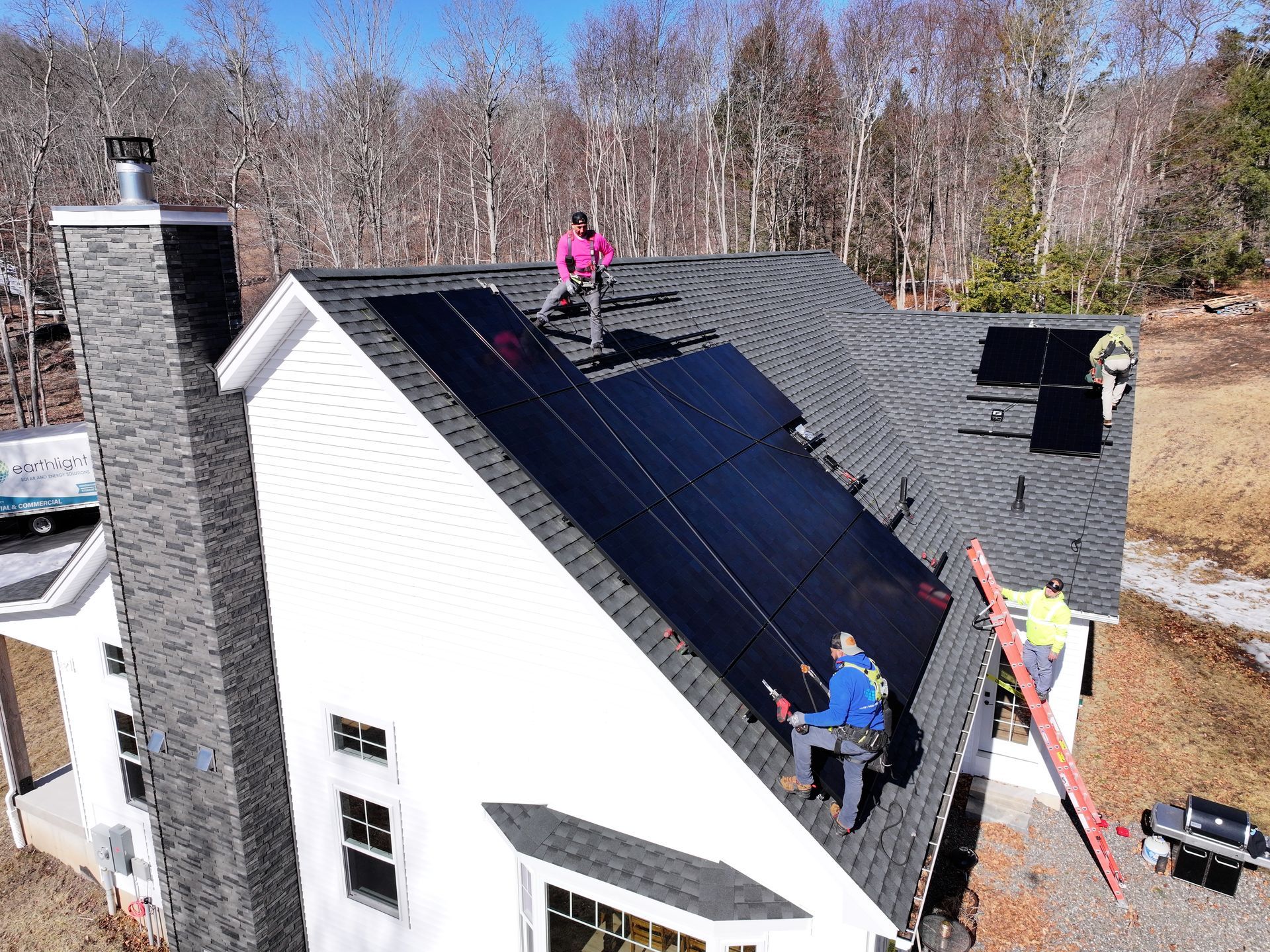 Workers installing dark roof panels on a white house roof, with ladders and tools.
