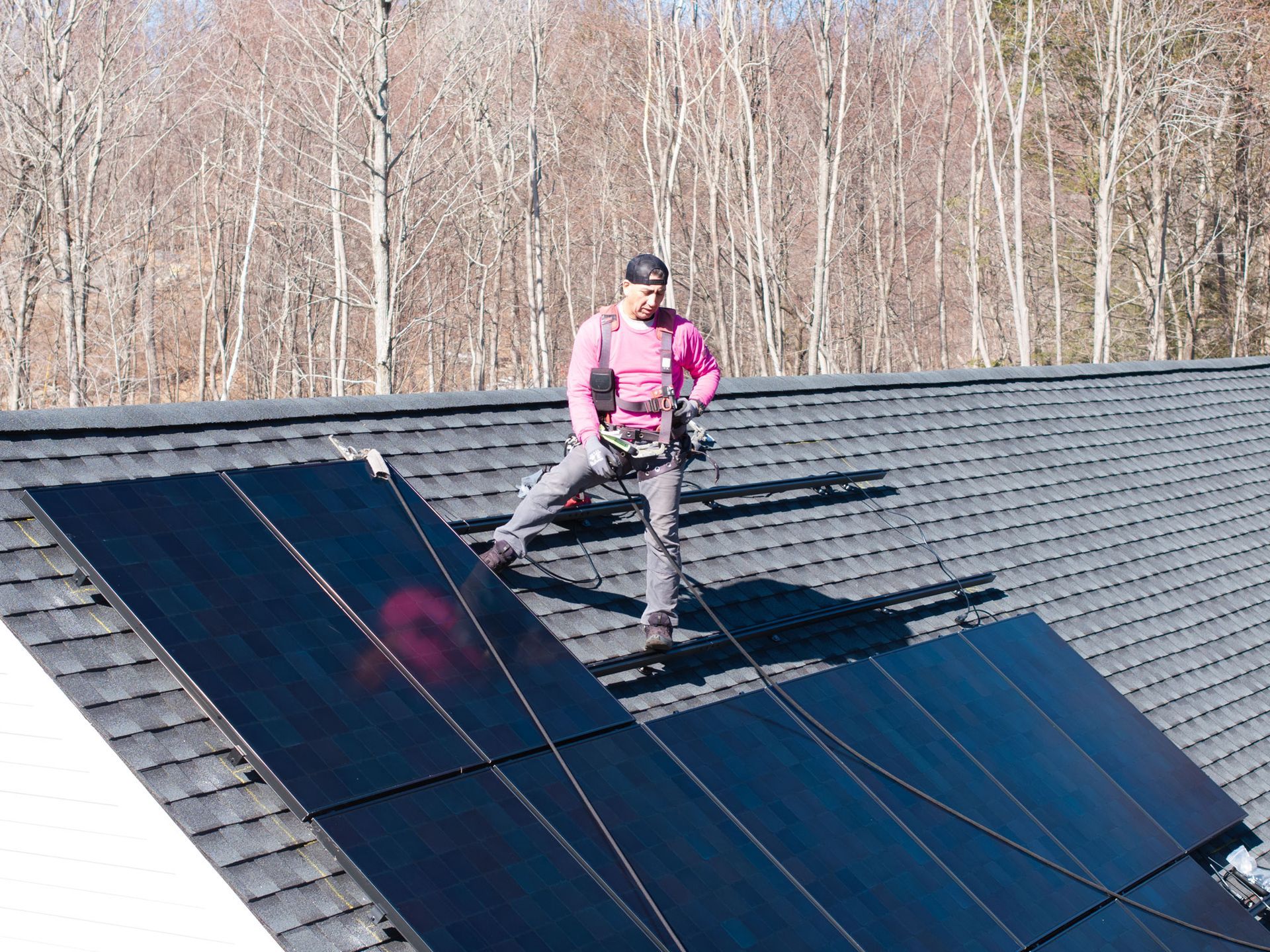 Worker standing on a rooftop beside black solar panels, wearing a pink jacket and safety gear.