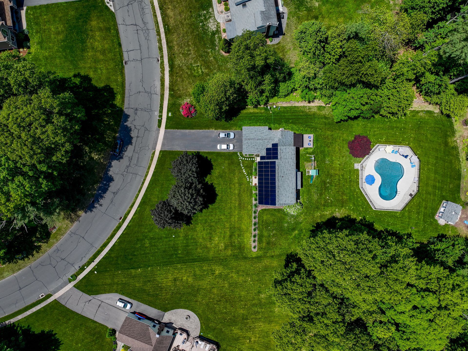 Aerial view of a house with solar panels, driveway, lawn, and a swimming pool beside a curved road.