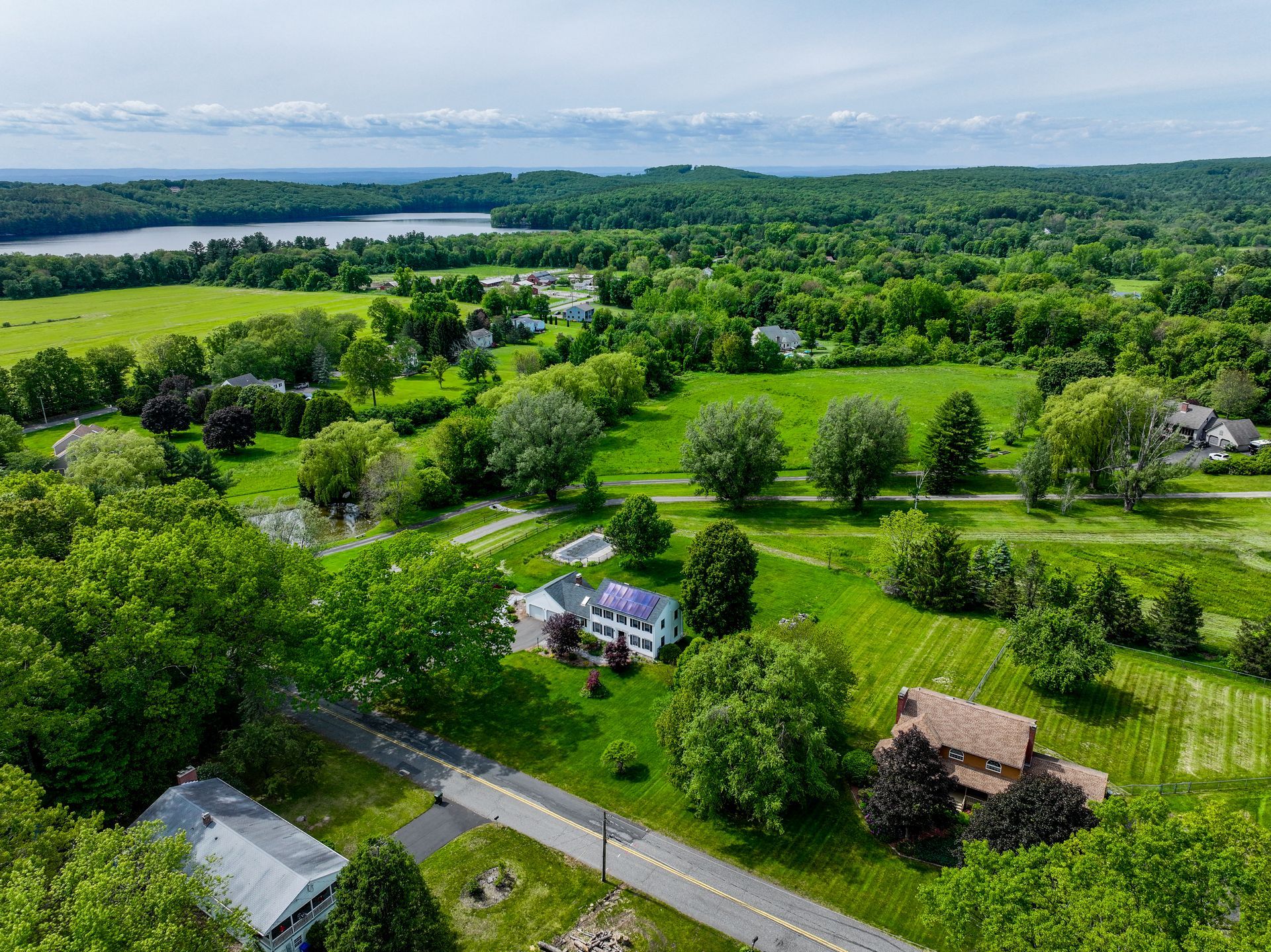 Aerial view of a green rural neighborhood with scattered houses, roads, and distant water under a cloudy sky