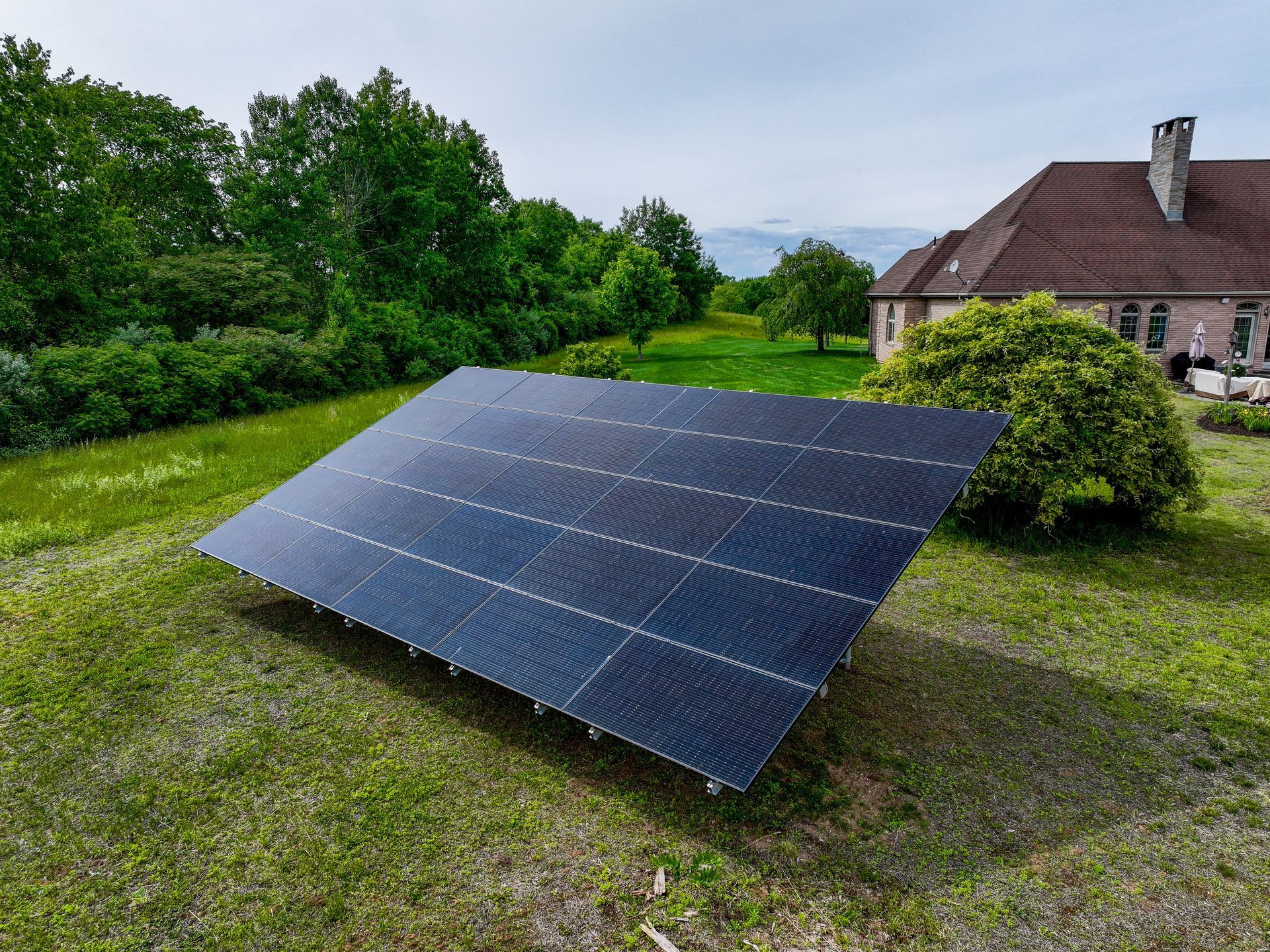 Solar panels installed on a grassy lawn near a house and trees on an overcast day