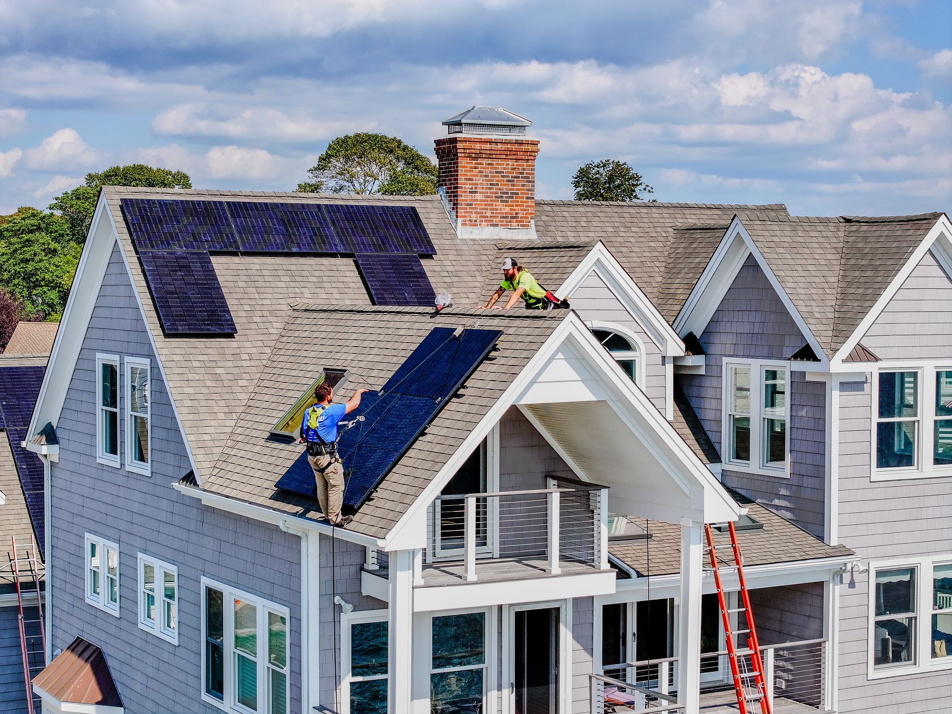 Workers installing solar panels on a gray house roof under a cloudy sky