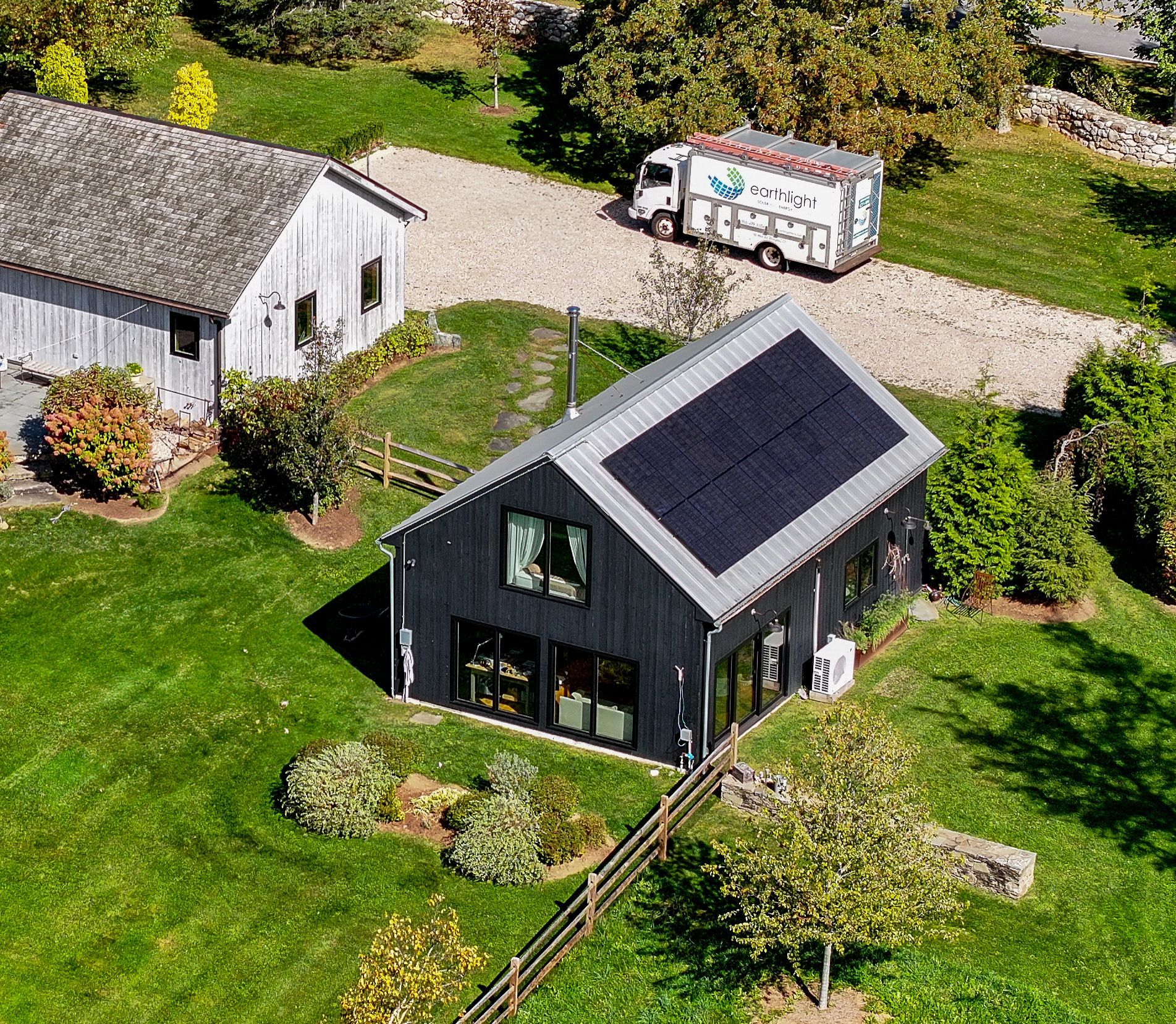 Aerial view of a black house with solar panels beside a white outbuilding and a van on a gravel drive.