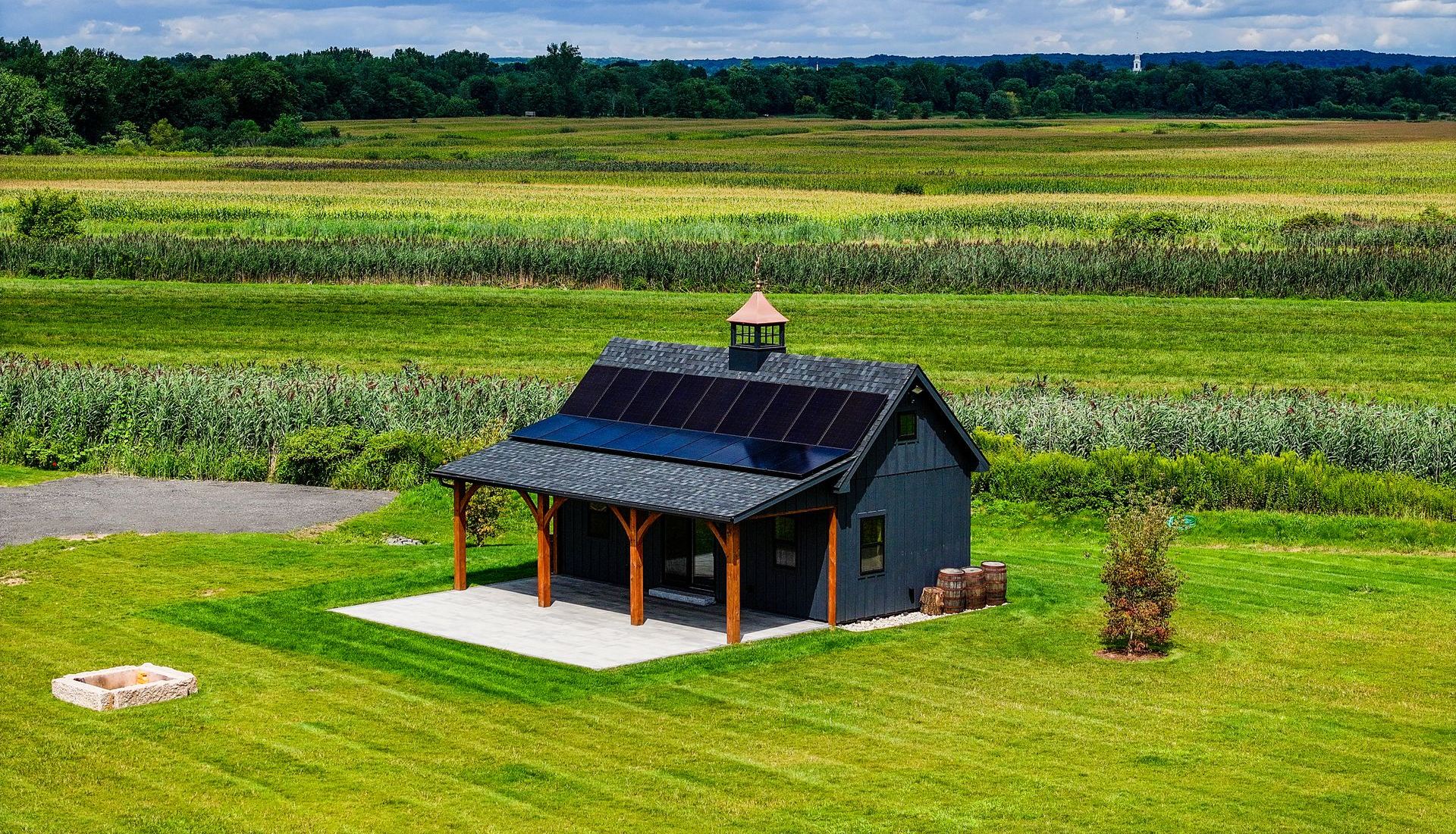 Small dark house with solar panels in a green field, surrounded by farmland and trees.
