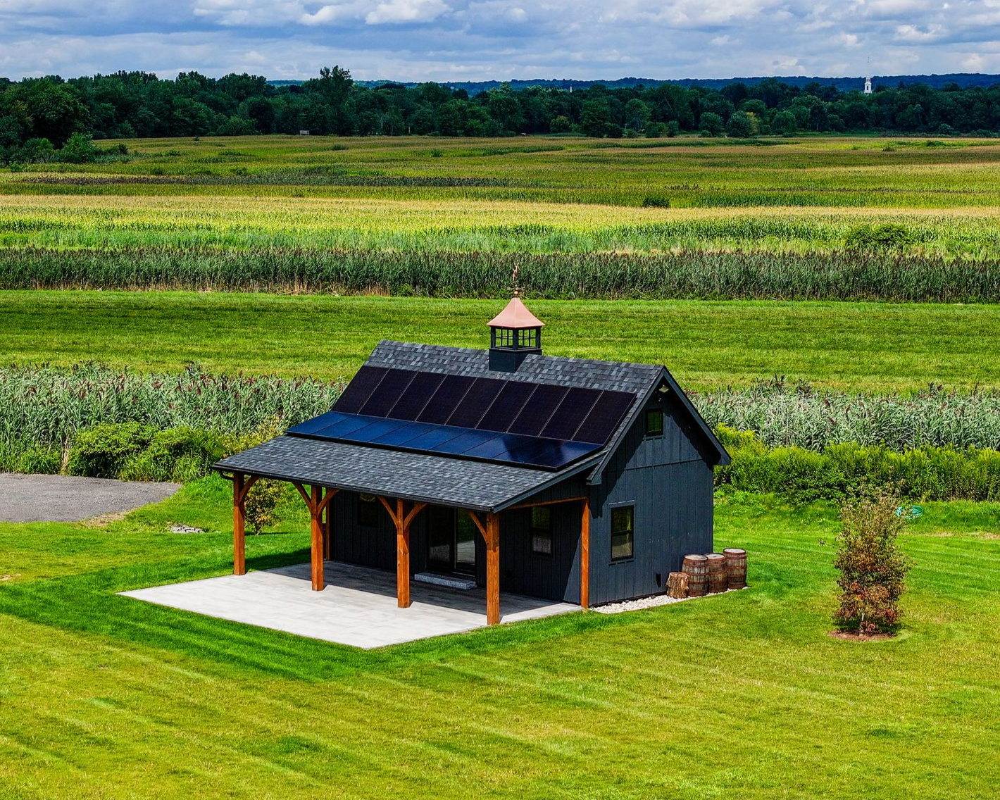Small dark-roofed shed with solar panels in a green field, bordered by trees and farmland.