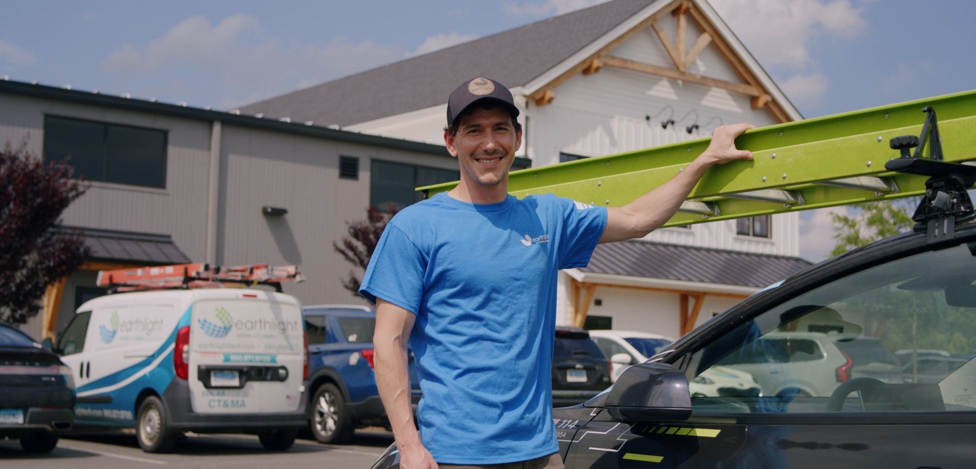 Smiling man in blue shirt standing by a car with a green kayak on top in a suburban driveway