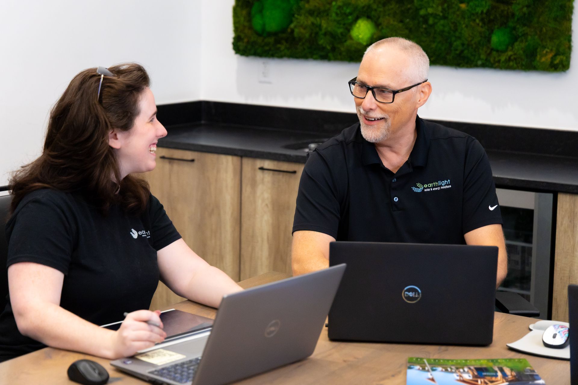 Two coworkers in black shirts talking at a table with laptops in a modern office