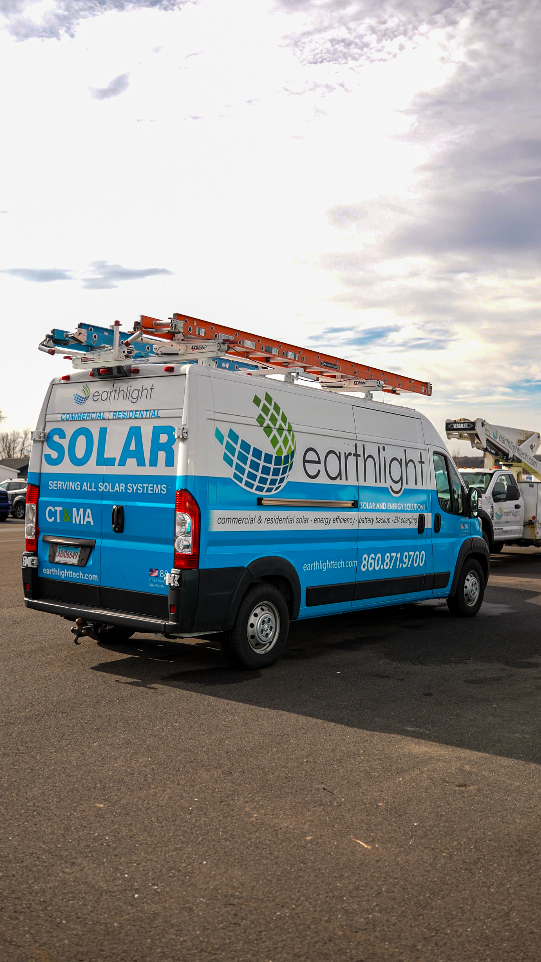 Blue utility van with solar panels and earth logo parked outdoors on a road under a cloudy sky