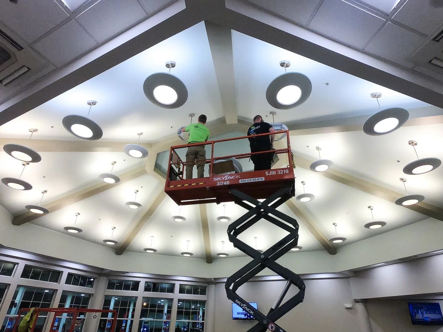 Two people on a red scissor lift near a bright ceiling with circular lights in a lobby.