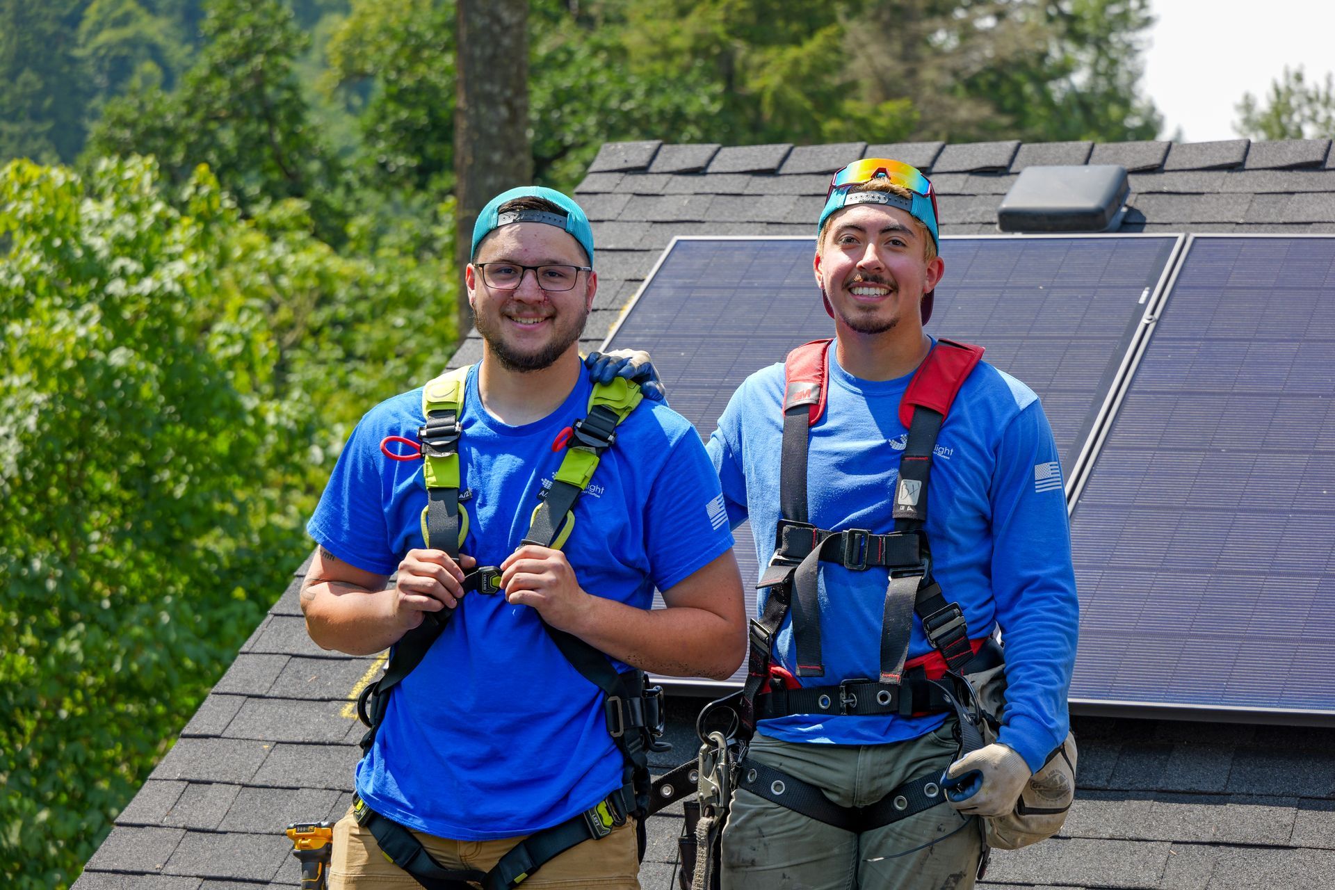 Two smiling workers in blue shirts and safety harnesses standing on a rooftop with solar panels