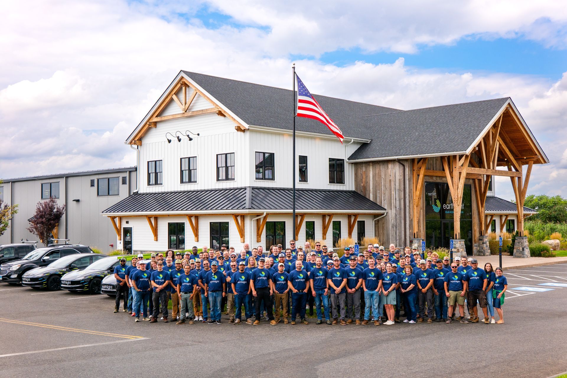 Large group posing in front of a white lodge with an American flag and parked cars