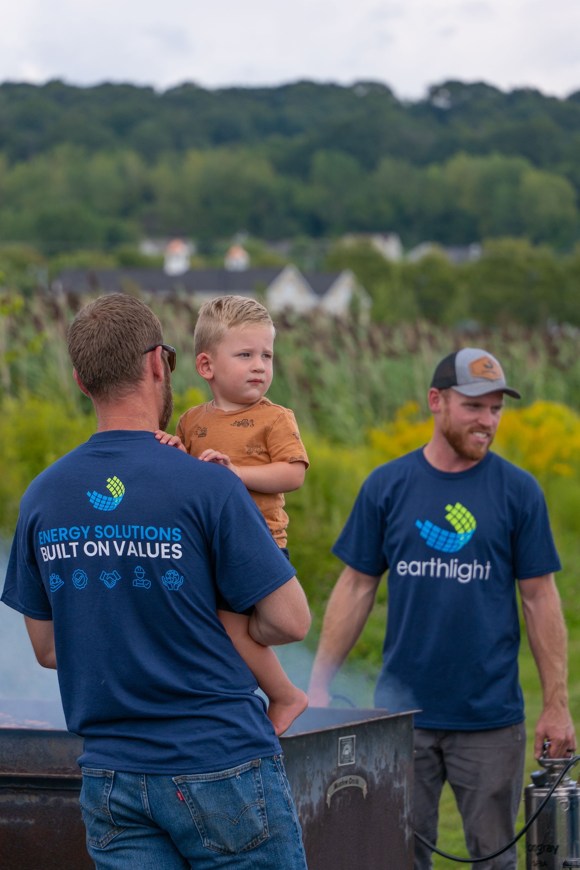 Two men in blue Earthlight shirts stand outdoors, one holding a toddler near a planting bed.