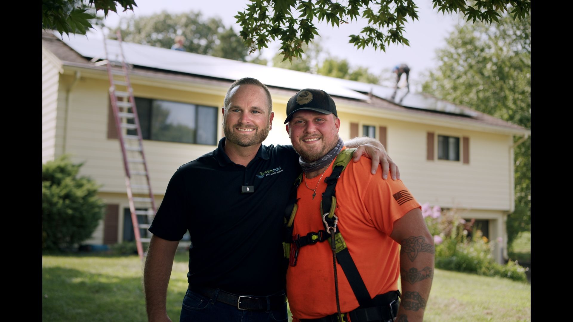 Two smiling workers standing in a yard in front of a beige house, one in black and one in orange safety gear