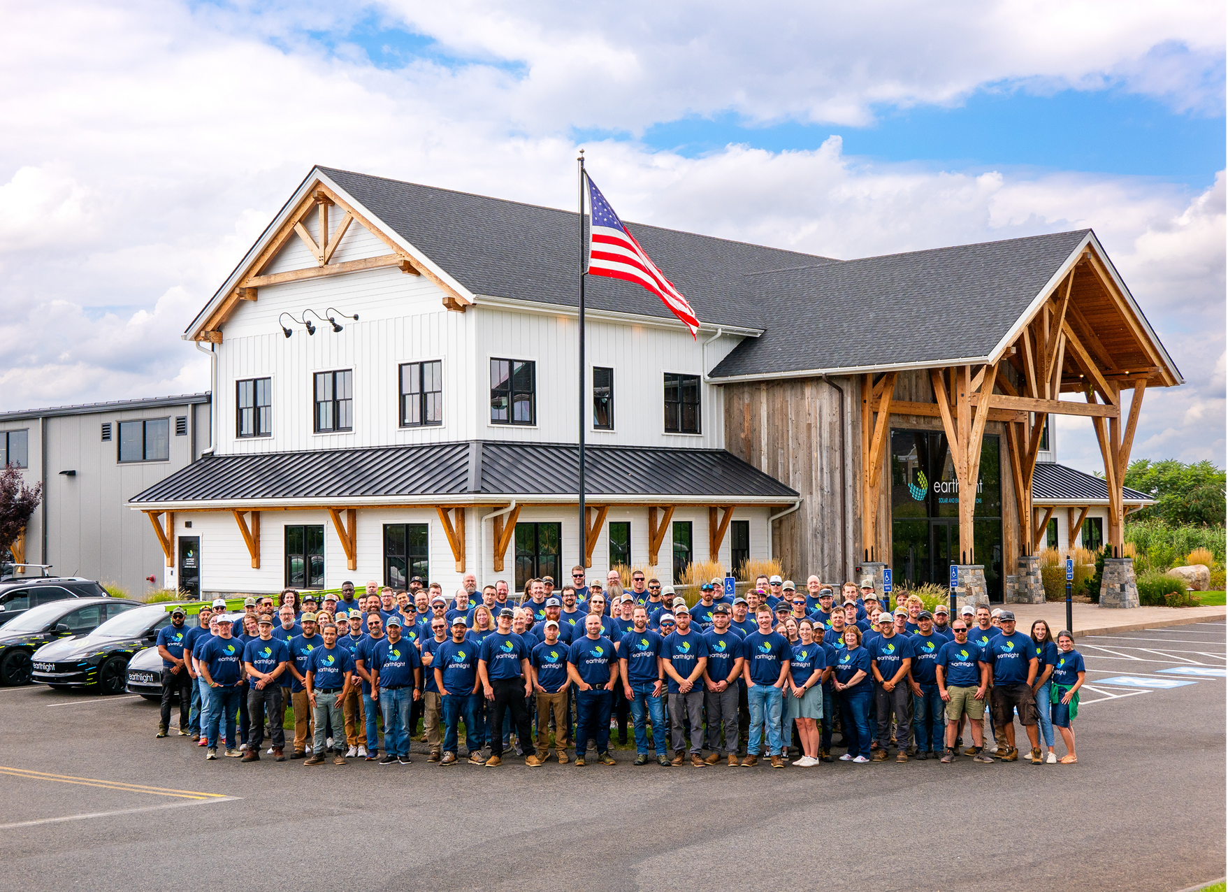 Large group posing outside a white lodge with a U.S. flag and parked cars in front