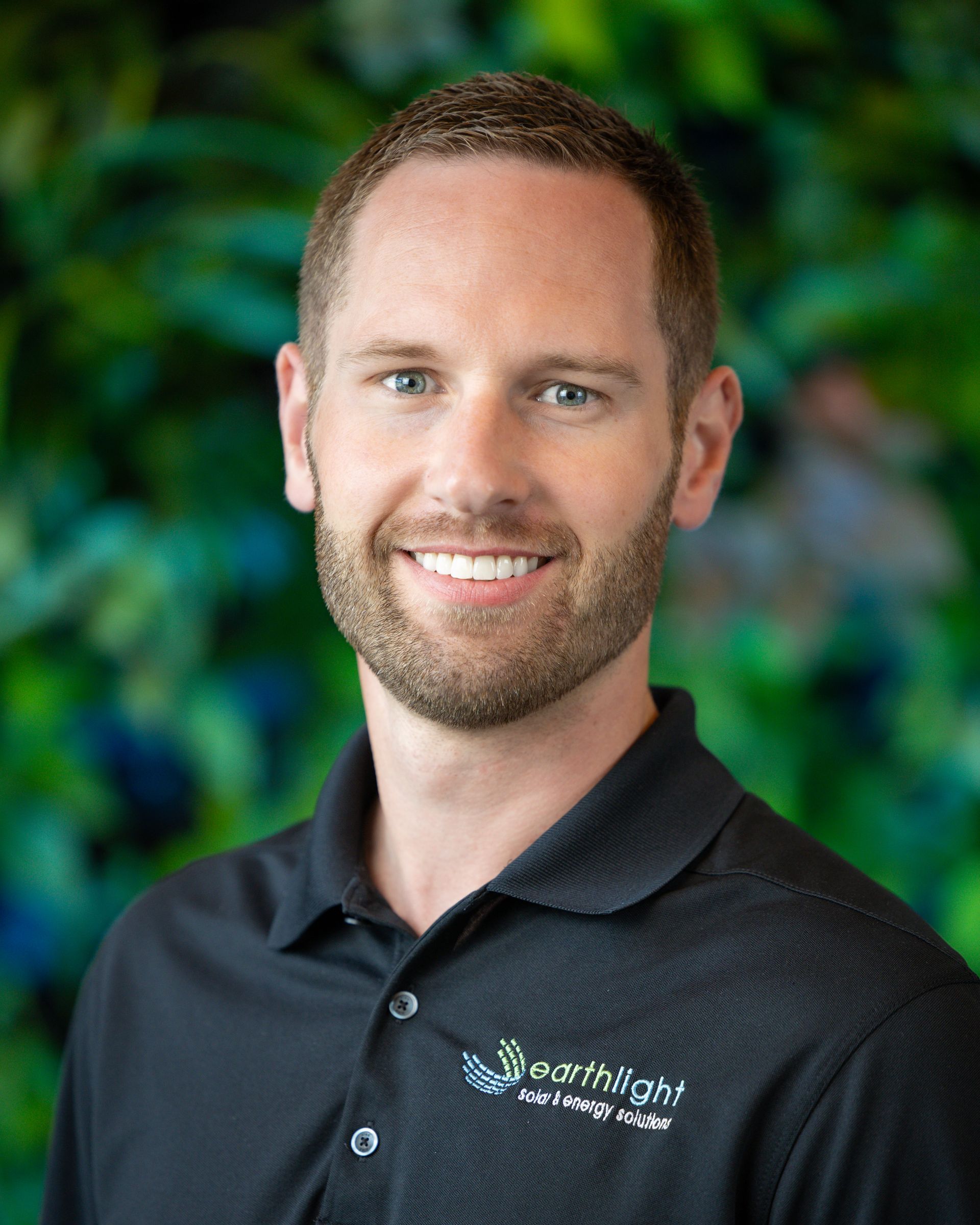 Smiling man in a black polo shirt with a logo, standing against a blurred green background