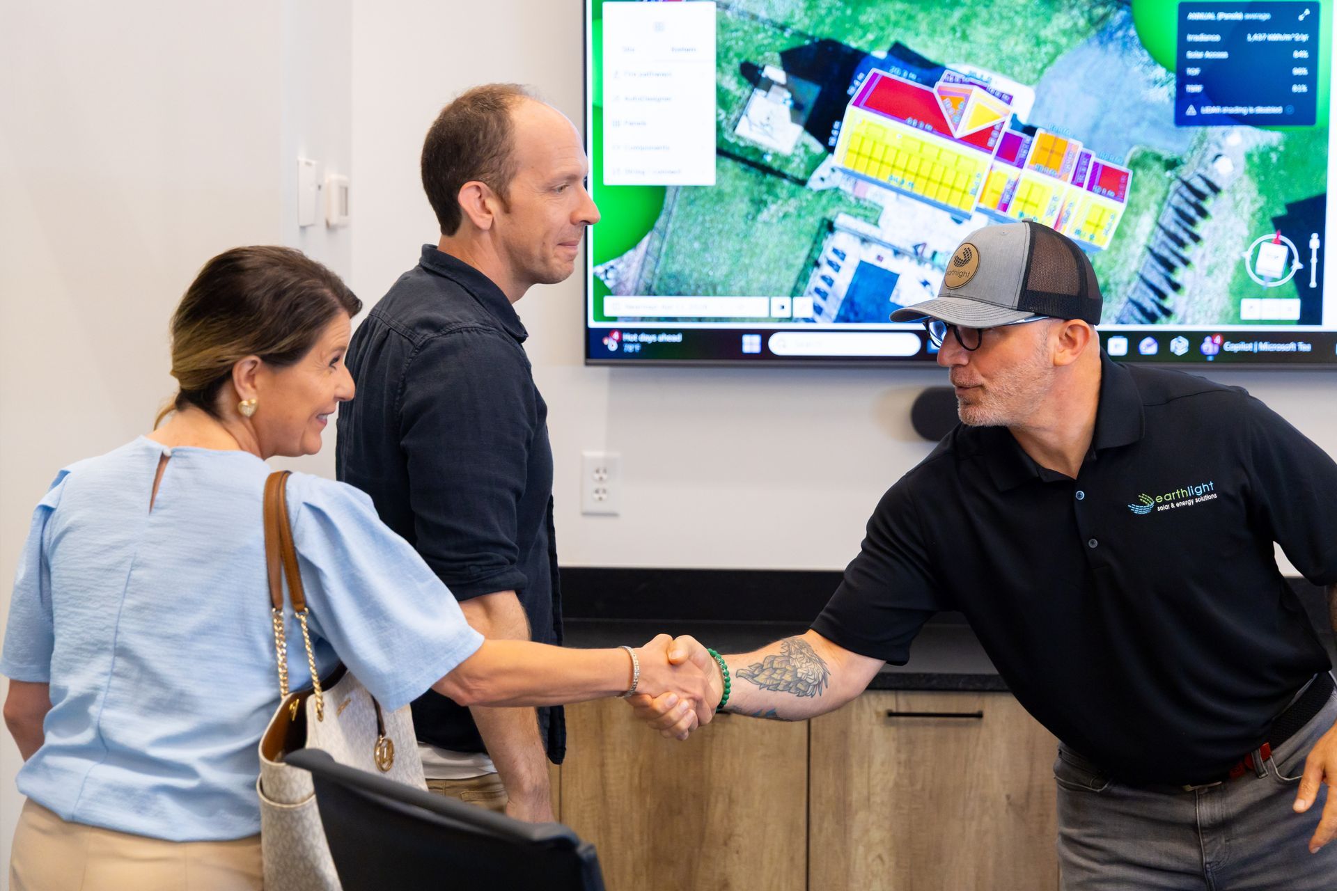 Three people shake hands in an office while a large display shows a site plan in the background.
