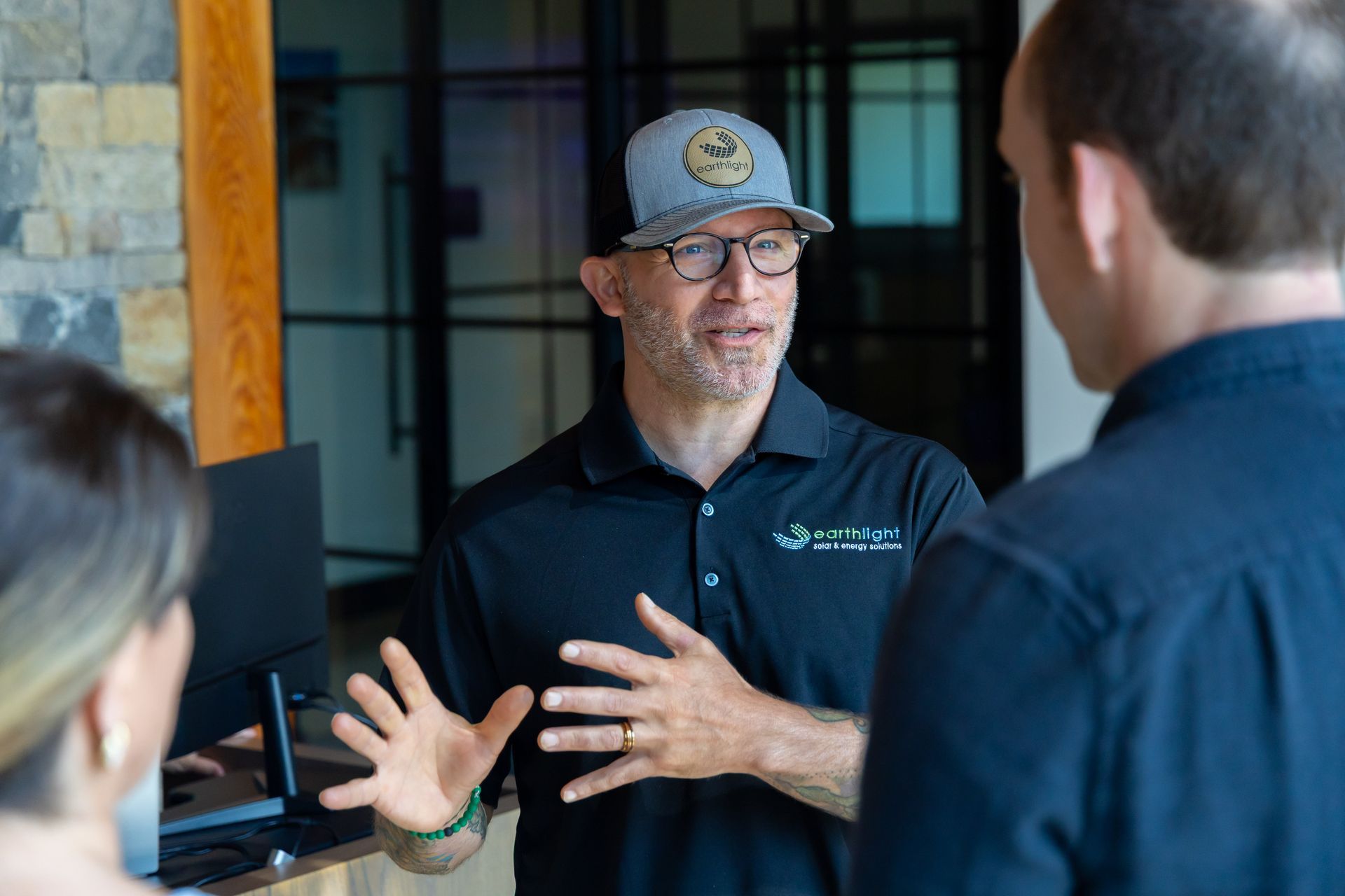 Man in black polo and cap speaking with two people indoors, hands gesturing.