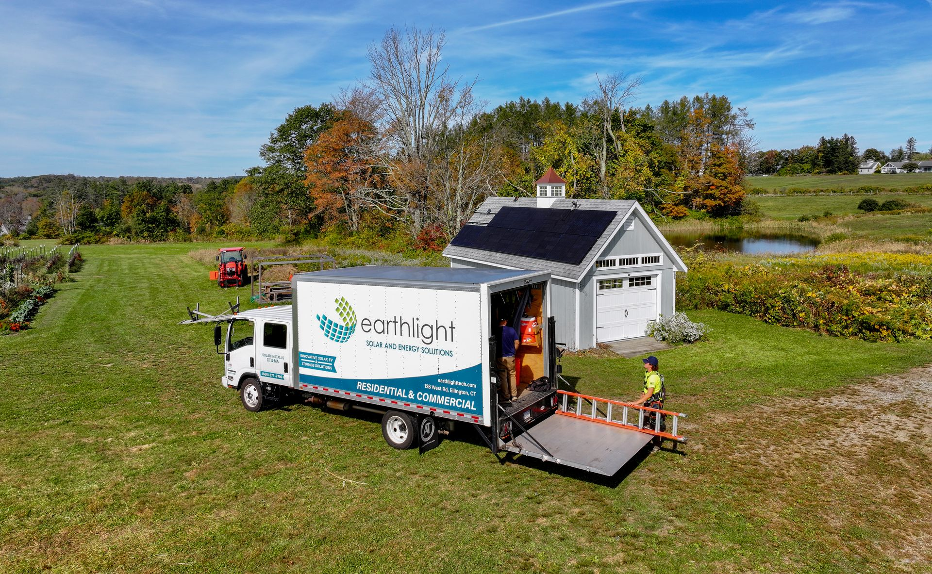 White delivery truck with an open rear ramp parked beside a small house in a green rural field.