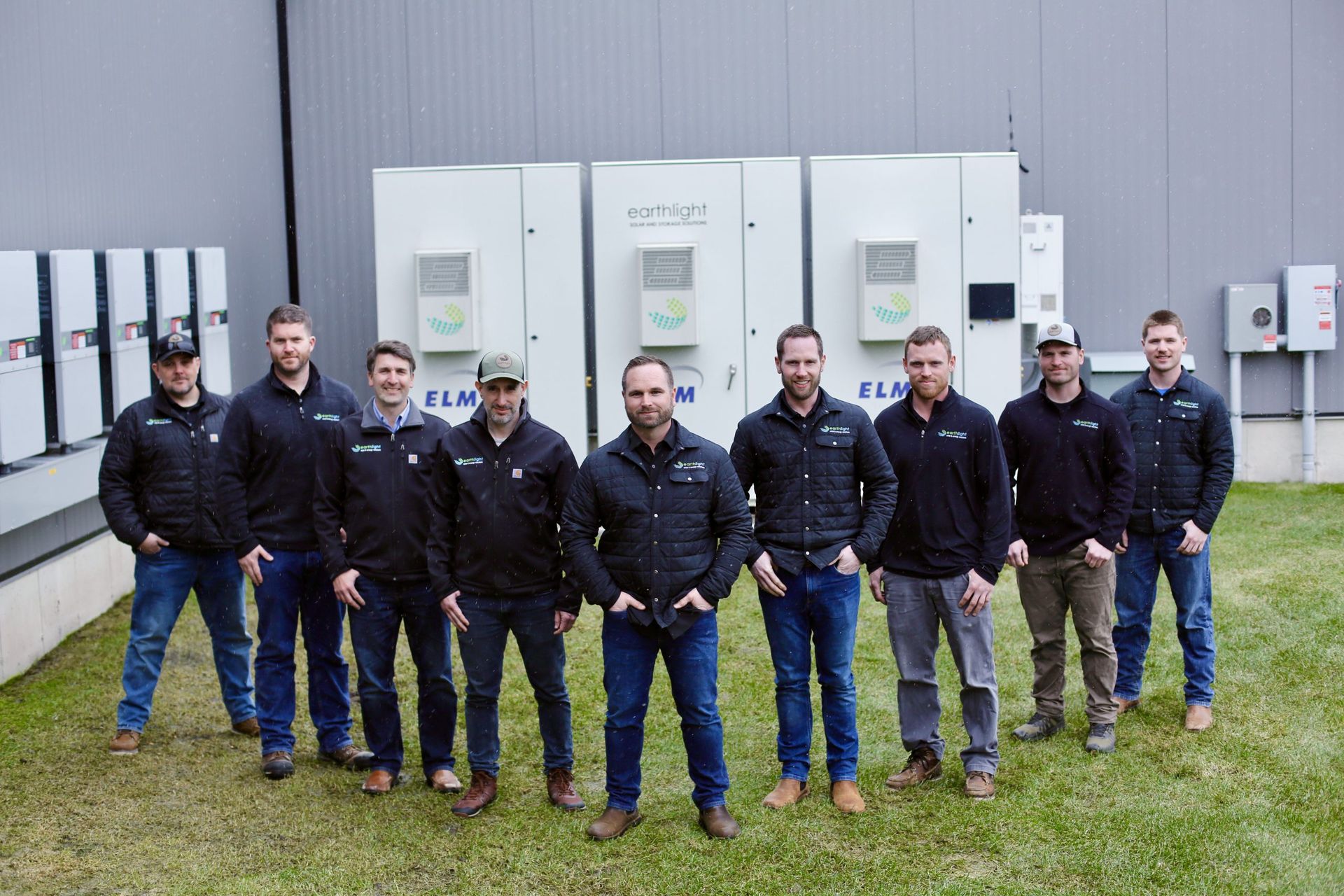 Group of people standing in front of large industrial units, wearing dark jackets on a grassy field