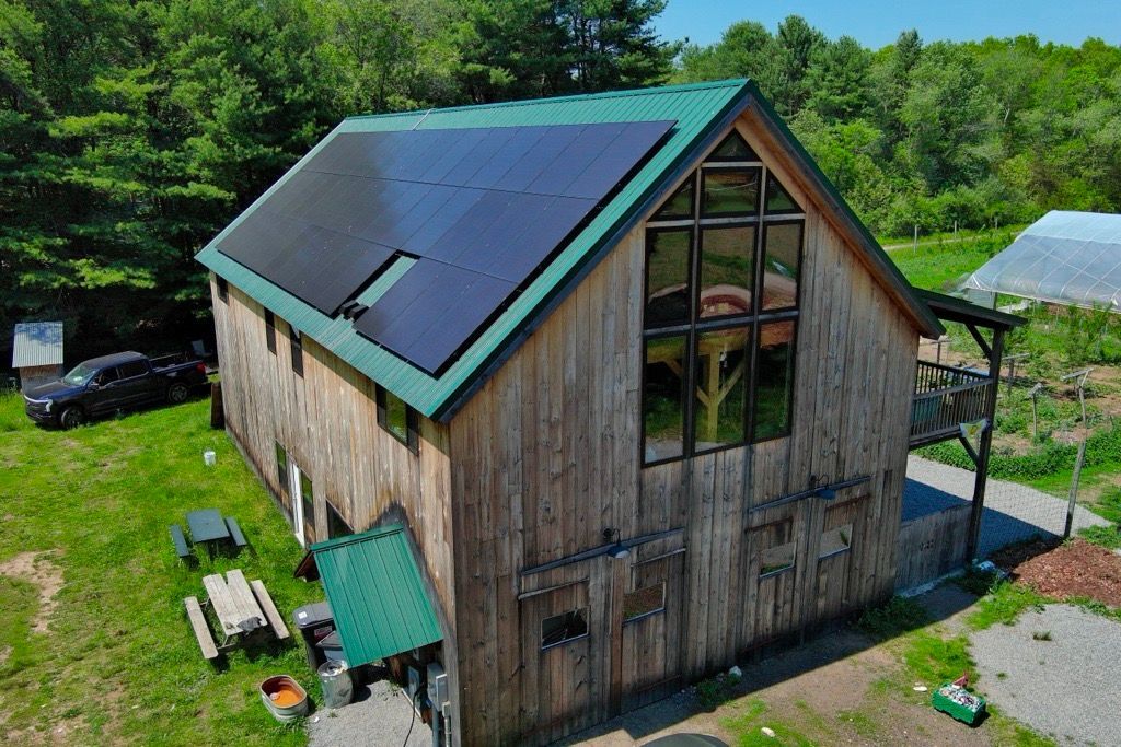 A rustic wooden barn with a dark roof and solar panels, surrounded by trees and a gravel path.