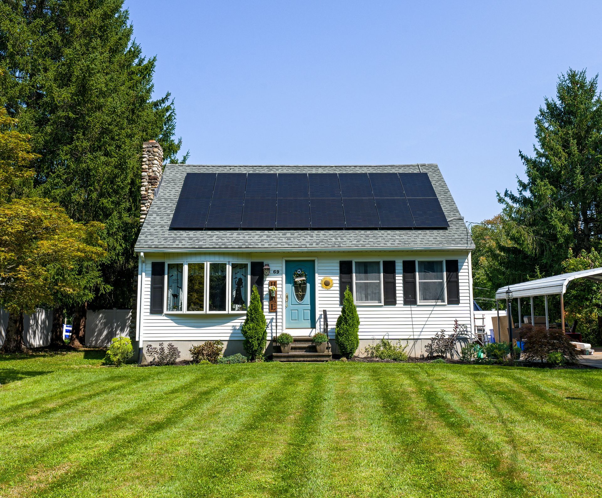 Small white house with solar panels, green lawn, and trees under a clear blue sky