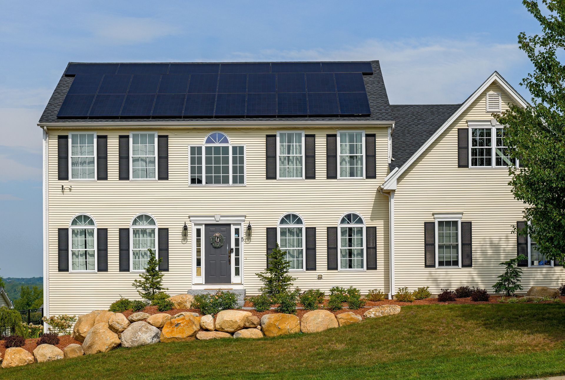 Large white two-story house with black shutters and solar panels on the roof, fronted by a landscaped lawn.