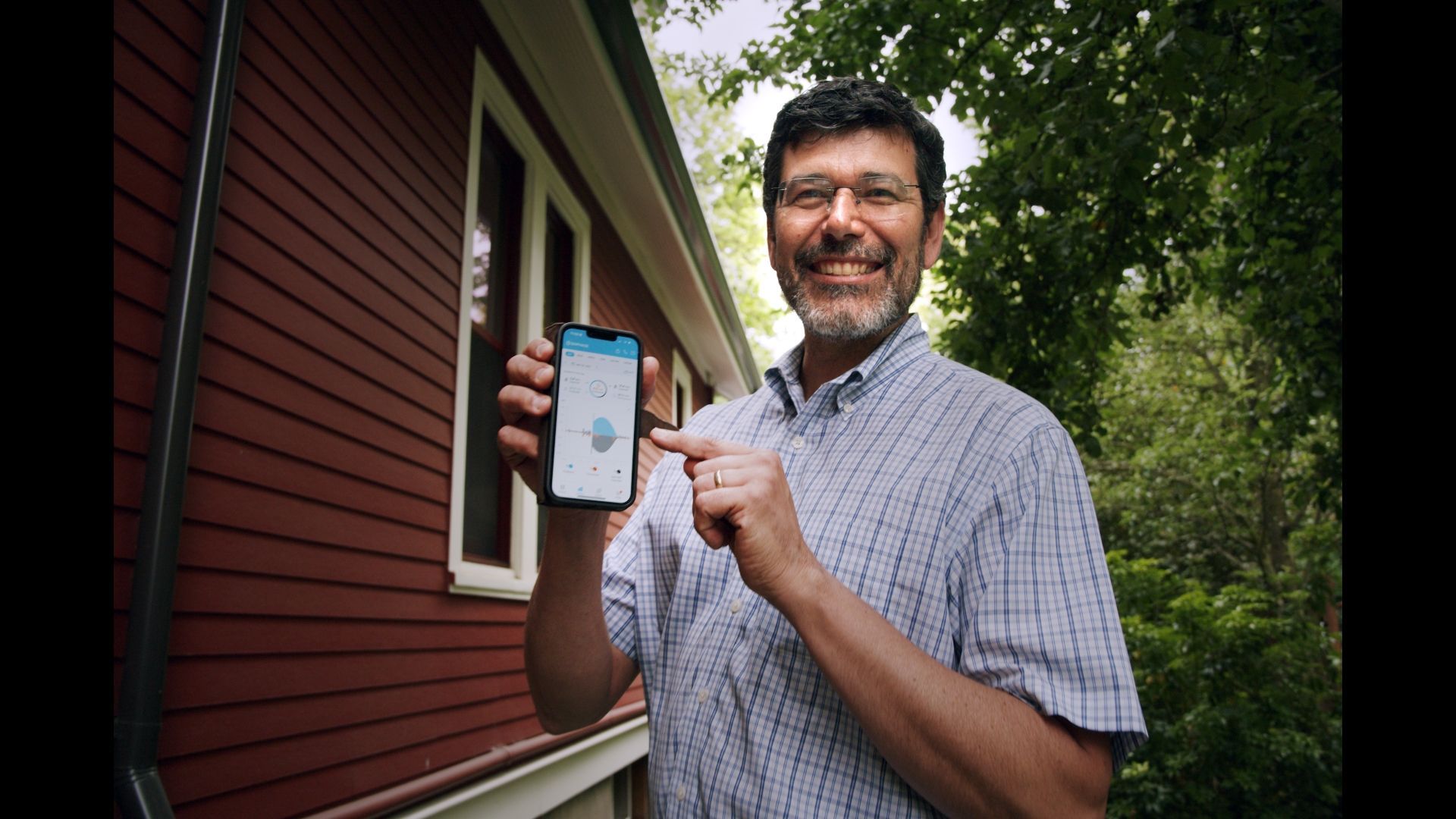 Man smiling outdoors beside a house, holding up a smartphone with an app on the screen.