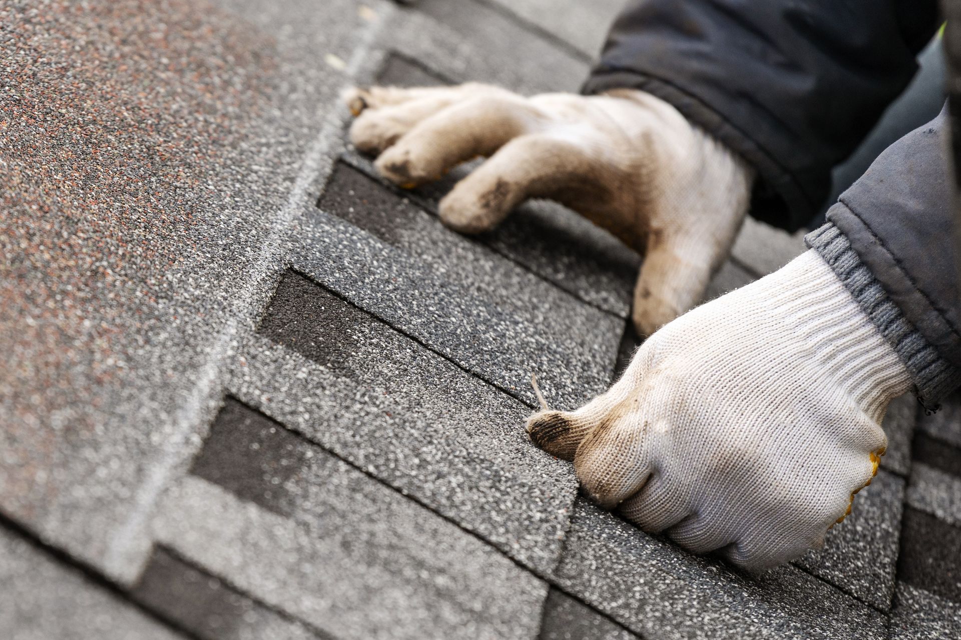 Hands in work gloves repairing gray roof shingles with a tool