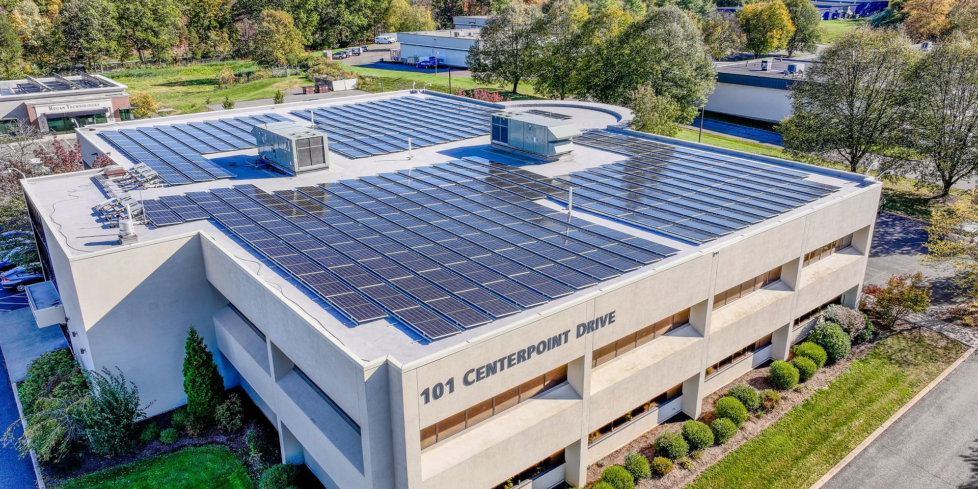Aerial view of a modern building with solar panels on the roof and surrounding trees.