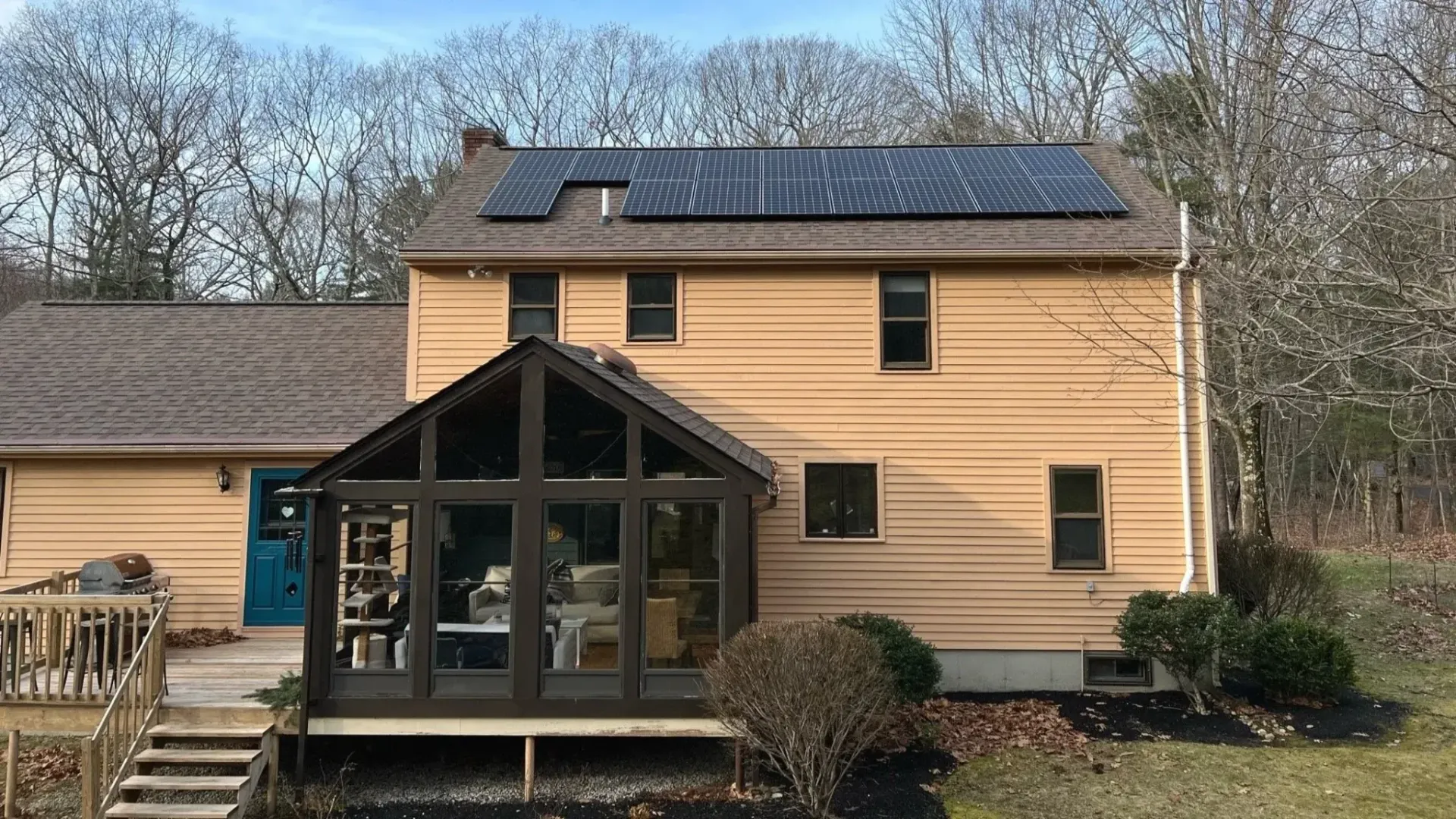Two-story tan house with a glass sunroom and solar panels, viewed from the backyard.