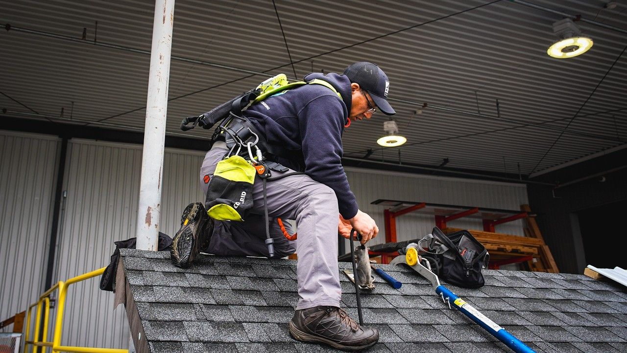 Worker in safety gear climbing a steep roof with tools at a building worksite.